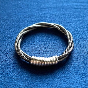 Puede incluir: Una pulsera plateada compuesta por hebras de metal enrolladas. La pulsera presenta una serie de pequeñas cuentas redondeadas agrupadas en un punto, agregando un elemento decorativo. La pulsera se muestra sobre un fondo azul.