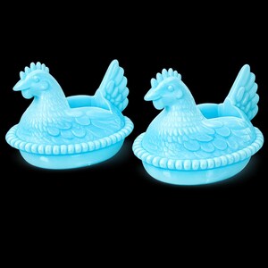 May include: Two light blue chicken-shaped egg holders. Each hen is sitting in a basket-like base. The hens have detailed feathers, a comb, and a beak. The background is black.