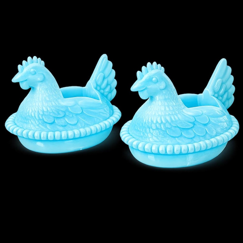 May include: Two light blue chicken-shaped egg holders. Each hen is sitting in a basket-like base. The hens have detailed feathers, a comb, and a beak. The background is black.
