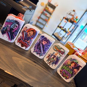 Peut inclure: Cinq étuis rectangulaires blancs en plastique pour vapoteuses, ornés d'impressions graphiques colorées de Michael Myers, Chucky, Rumi, Dragon Ball Z et Hocus Pocus. Chaque étui présente des illustrations détaillées avec des logos textuels.