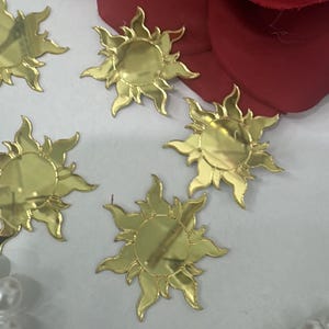 May include: Gold-colored sun-shaped embellishments with a reflective surface. The suns have pointed rays and a circular center. The image also shows a red fabric and a string of white pearls.