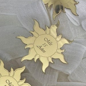May include: Gold-colored sun-shaped decorative pins. Each pin has a central circle with the inscription "Mis 15 Años". The pins are arranged on a white fabric background, with the sun's rays extending outwards.
