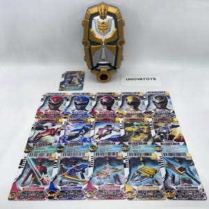May include: A gold and silver shield-shaped toy with a red button, and a collection of colorful trading cards. The cards feature images of characters and vehicles. The text "UNOVATOYS" is visible.