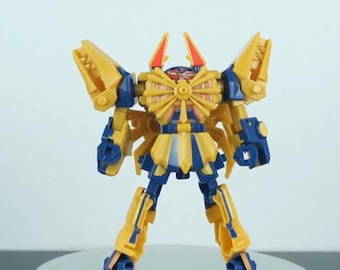 May include: A yellow and blue toy robot with articulated limbs and a detailed design. The robot has a central body with a circular design, two red horns, and large yellow wings. The toy is standing on a silver surface.