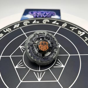 Peut inclure: Un toupie grise et noire avec un motif de tigre orange au centre. Le jouet est posé sur une surface noire avec des formes géométriques blanches. Le logo Unova Toys est visible en arrière-plan.