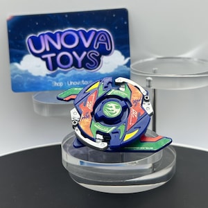 May include: A blue, green, and orange Beyblade spinning top toy featuring a central green emblem with a bird graphic and the text Dranzer. The toy has metallic silver accents and is displayed on a clear stand with a sign reading Unova Toys.