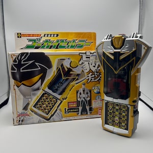 Peut inclure: Un appareil jouet jaune et argenté avec un écran rabattable, à côté d'une boîte avec du texte japonais et une illustration de personnage. L'appareil a un panneau de boutons et un design en forme de bouclier. La boîte présente un personnage avec un casque noir et argenté.