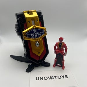 May include: A black, red, and gold toy with the text "GOKAIGER" and a small red action figure. The toy has a shield-like design with a gold center and a black base. The action figure is red with black and gold accents.