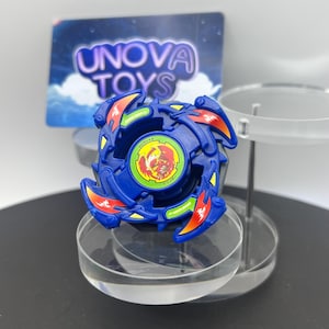 May include: A blue plastic spinning top toy featuring four curved blades with orange and green accents. The center includes a circular emblem with a red phoenix graphic and Japanese text. The toy is displayed on a clear circular stand.