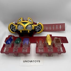 May include: A red and gold toy belt with several coloured accessories. The belt buckle has a bat-like design. The accessories are various colours, including blue, green, purple, and gold. The text "UNOVATOYS" is visible on a white label.