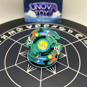 May include: Green and blue spinning top toy with red and yellow accents. The toy has the word "DRIGER" printed on it. The background includes a box with the words "UNOVA TOYS".