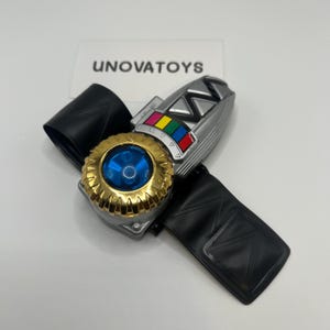 Puede incluir: Una pulsera de juguete plateada y negra con un dial circular dorado con un centro azul. La pulsera tiene una correa negra y una sección con botones coloridos. El texto "UNOVATOYS" es visible en el fondo.