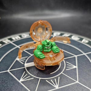 May include: A translucent orange and green toy figure with articulated parts. The figure has a round base and a hinged top, with a green humanoid figure inside. The toy is set against a patterned background.