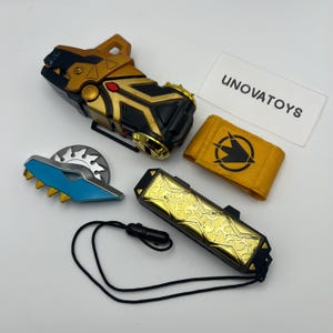 May include: A set of toy accessories, featuring a gold and black device with a dog-like head, a blue and silver saw-like piece, a gold and black rectangular device, and a yellow armband with a black symbol. The text "UNOVATOYS" is visible.