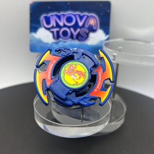 May include: A blue spinning top toy featuring orange and yellow flame-like accents on the outer ring. The center displays a circular green sticker with a red phoenix graphic and the text Dranzer-S. A blue card with Unova Toys text is behind.
