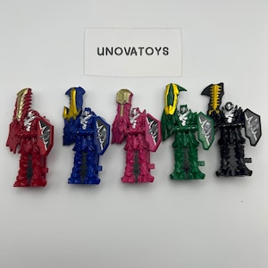 Puede incluir: Cinco juguetes de figuras de acción coloridas en rojo, azul, rosa, verde y negro. Cada figura tiene un arma y un escudo únicos. Los juguetes están dispuestos en fila sobre un fondo blanco. El texto "UNOVATOYS" es visible.