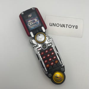 Puede incluir: Un teléfono de juguete plateado y rojo con teclado y pantalla. La pantalla muestra la palabra "CHANGE" y otro texto. El teclado tiene botones numéricos rojos. El teléfono está parcialmente abierto.