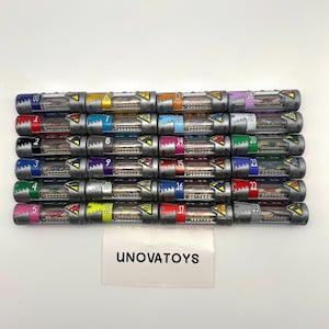 Puede incluir: Una colección de veinticuatro cápsulas de juguete cilíndricas, cada una con un cuerpo plateado y una parte superior de color. Las cápsulas están numeradas del 00 al 23 y contienen pequeñas figuras detalladas. El texto "UNOVATOYS" es visible.