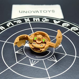 May include: A tan toy with a hinged design, featuring green and brown accents. The toy is open, revealing internal components. The background includes a black surface with white geometric patterns and the text "UNOVATOYS".