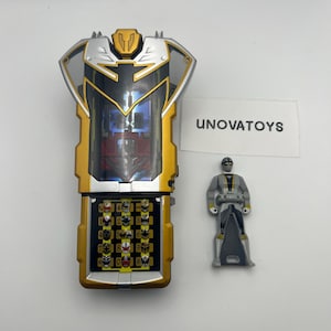 Peut inclure: Un jouet doré et argenté avec un clavier et une petite figurine. Le jouet a une forme rectangulaire avec un fond articulé et une fenêtre transparente. La figurine est grise et argentée avec un casque noir.