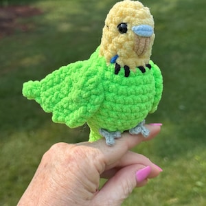 May include: A handmade crochet parakeet in bright green and yellow. The bird has black eyes, a blue patch, and a tan beak. The parakeet is perched on a finger, showcasing its detailed craftsmanship and vibrant colours.