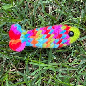 Peut inclure: Un petit poisson en crochet fait main avec un motif en laine multicolore aux tons rose, bleu, orange, jaune et vert. Ce jouet présente un œil en perle de plastique noire et une nageoire caudale distincte, le tout réalisé en mailles texturées.