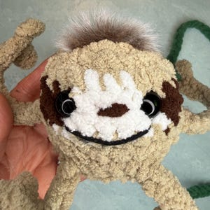 May include: A crocheted sloth-shaped bag made of tan yarn with a white face patch, dark brown eye patches, and black plastic safety eyes. It features a tuft of brown faux fur on top, a stitched black mouth, and a long green braided yarn strap.