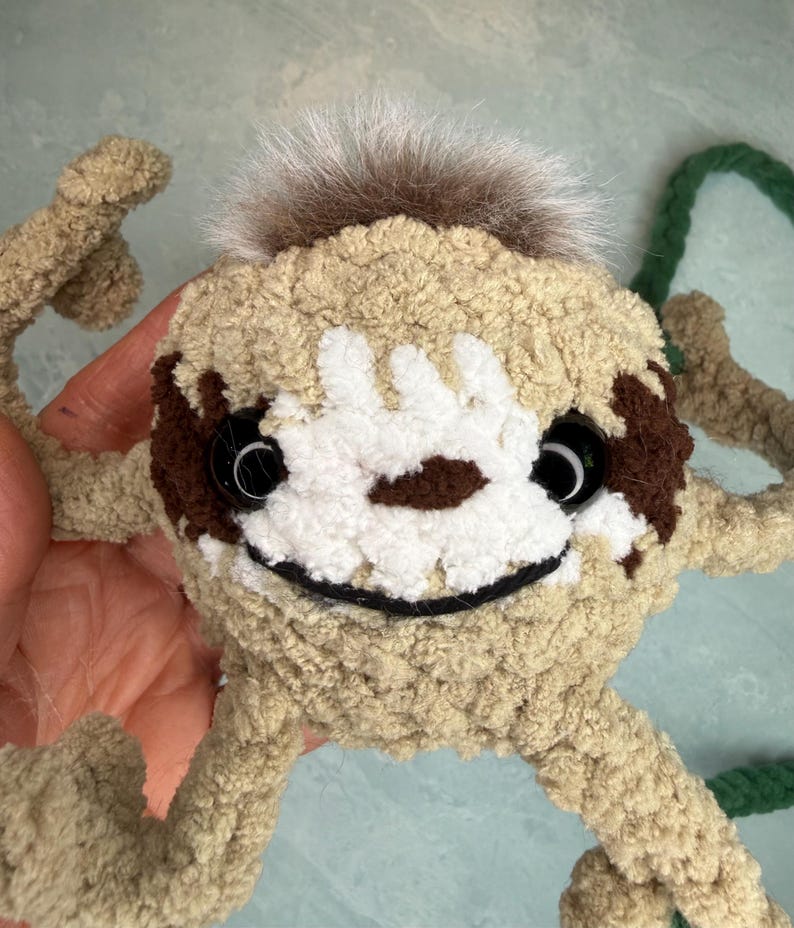 May include: A crocheted sloth-shaped bag made of tan yarn with a white face patch, dark brown eye patches, and black plastic safety eyes. It features a tuft of brown faux fur on top, a stitched black mouth, and a long green braided yarn strap.