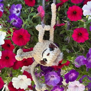 May include: A beige crocheted sloth plush toy with dark brown eye patches and black safety eyes, hanging by its arms from a grey yarn loop. The sloth has a soft, fuzzy texture and is positioned against a background of vibrant petunia flowers.