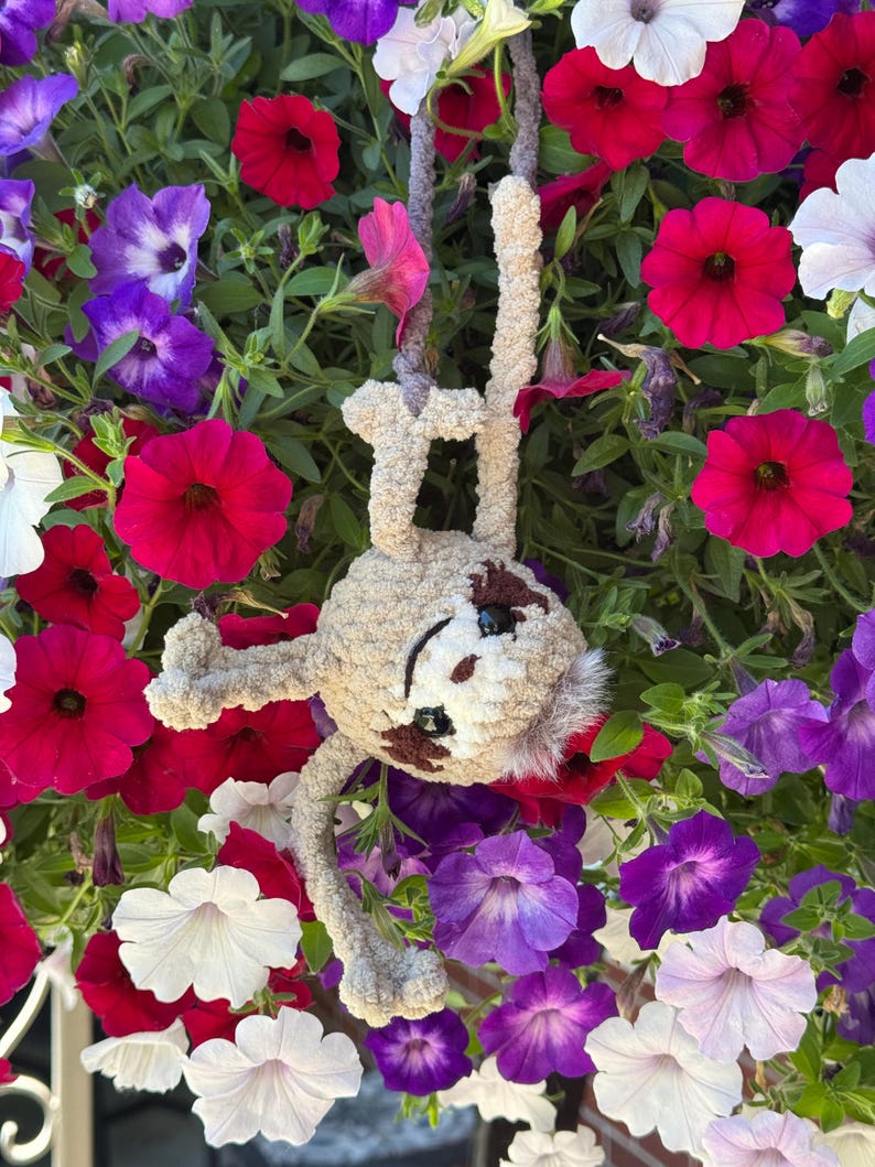 May include: A beige crocheted sloth plush toy with dark brown eye patches and black safety eyes, hanging by its arms from a grey yarn loop. The sloth has a soft, fuzzy texture and is positioned against a background of vibrant petunia flowers.