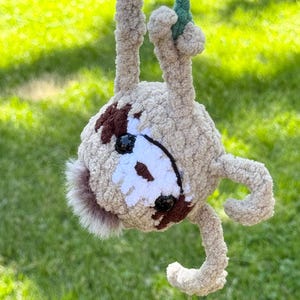 May include: A crocheted sloth plush toy made of light tan fuzzy yarn, hanging upside down from a green braided cord. The sloth features dark brown eye patches, a white face mask, black plastic eyes, and a small tuft of brown fur at the top.