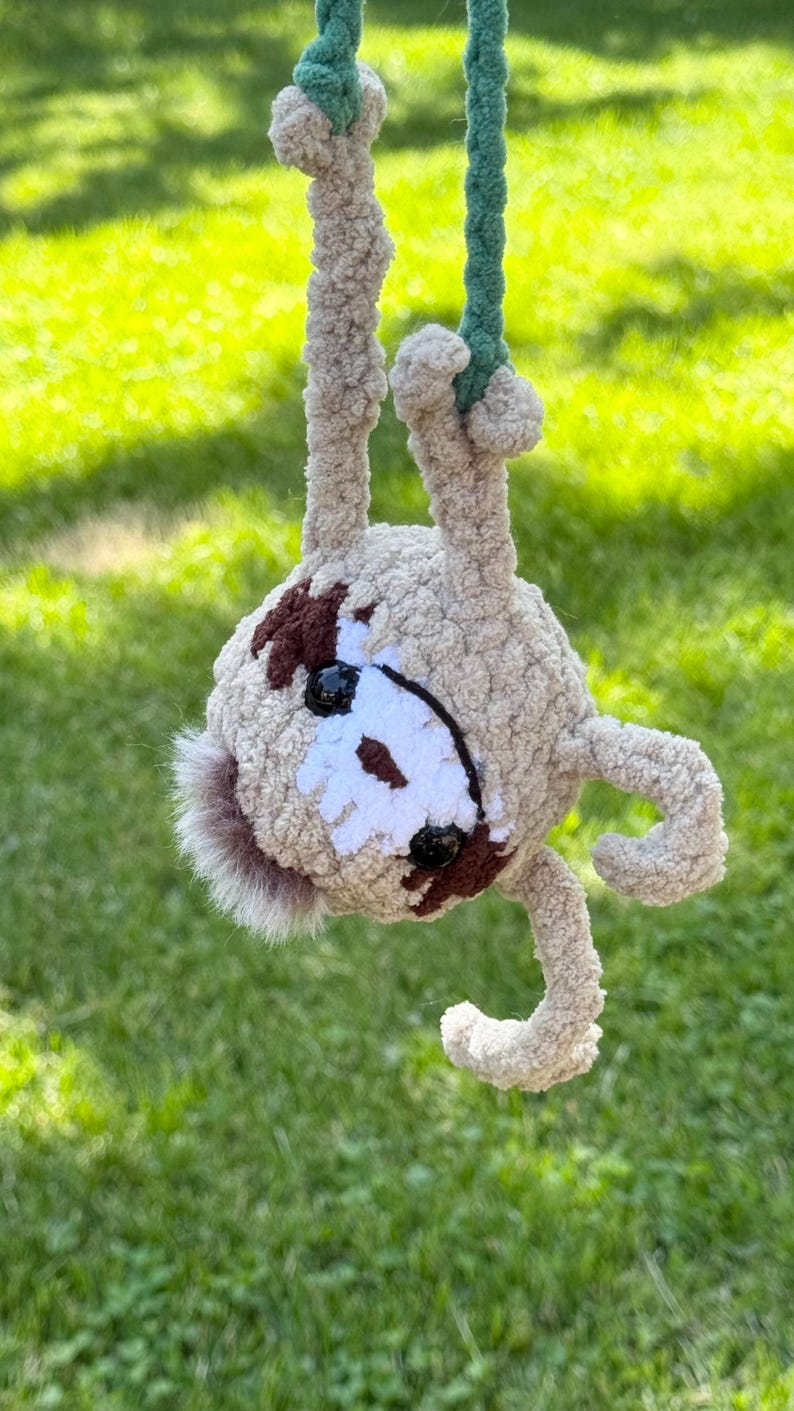May include: A crocheted sloth plush toy made of light tan fuzzy yarn, hanging upside down from a green braided cord. The sloth features dark brown eye patches, a white face mask, black plastic eyes, and a small tuft of brown fur at the top.