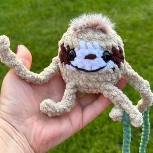 May include: A small, round, beige crocheted sloth plush toy with long, thin arms and legs. It features a white face patch, dark brown eye markings, black plastic eyes, a stitched black mouth, and a tuft of brown faux fur on the top of its head.