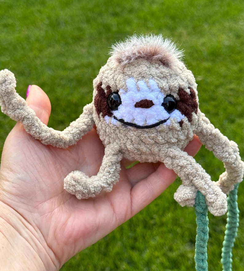 May include: A small, round, beige crocheted sloth plush toy with long, thin arms and legs. It features a white face patch, dark brown eye markings, black plastic eyes, a stitched black mouth, and a tuft of brown faux fur on the top of its head.