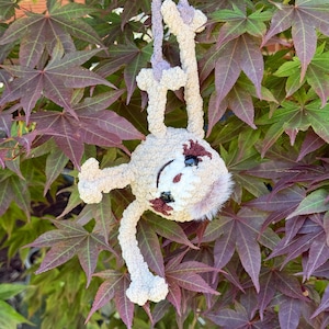 May include: A crocheted sloth plush toy made from soft, light beige yarn. The sloth has dark brown patches around its black plastic eyes, a white face, and a small tuft of light brown fur on its head. It hangs from a matching beige yarn loop.
