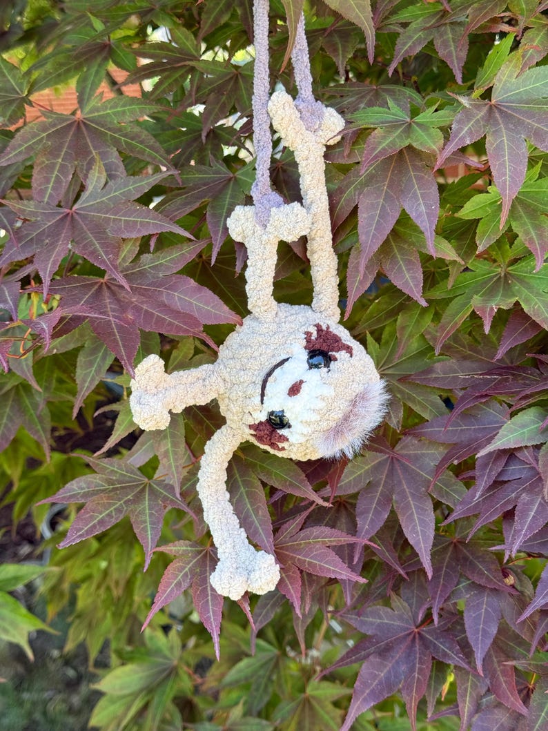 May include: A crocheted sloth plush toy made from soft, light beige yarn. The sloth has dark brown patches around its black plastic eyes, a white face, and a small tuft of light brown fur on its head. It hangs from a matching beige yarn loop.