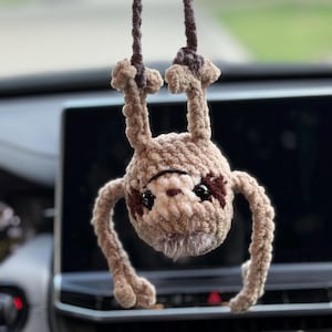 May include: A crocheted sloth hanging upside down by its feet from a dark brown yarn cord. The sloth is made of light brown plush yarn with dark brown patches around its black plastic eyes, a small brown nose, and a fuzzy white chin area.