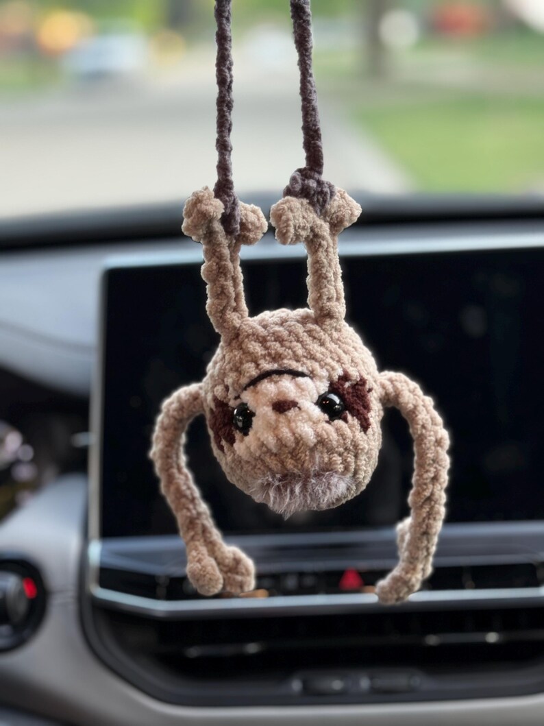 May include: A crocheted sloth hanging upside down by its feet from a dark brown yarn cord. The sloth is made of light brown plush yarn with dark brown patches around its black plastic eyes, a small brown nose, and a fuzzy white chin area.