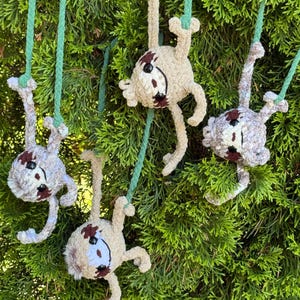 May include: Four crocheted sloth ornaments in beige yarn with dark brown facial markings and black eyes. Each sloth is suspended by a green braided cord, featuring articulated limbs and a soft, textured appearance, designed to hang from branches.