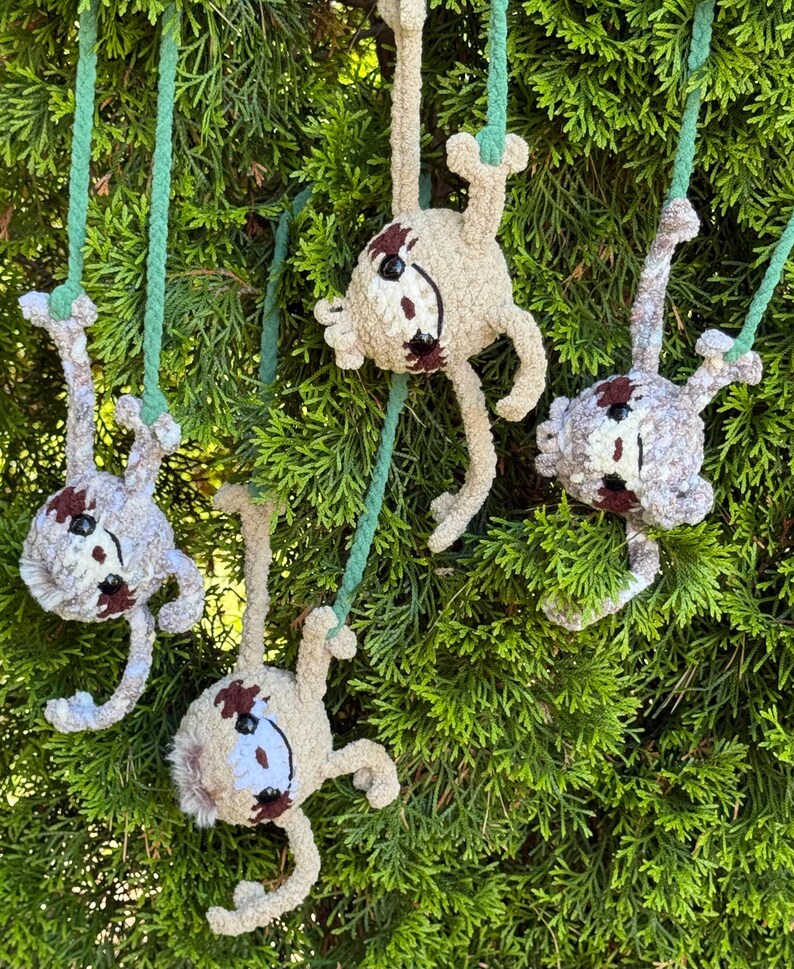 May include: Four crocheted sloth ornaments in beige yarn with dark brown facial markings and black eyes. Each sloth is suspended by a green braided cord, featuring articulated limbs and a soft, textured appearance, designed to hang from branches.