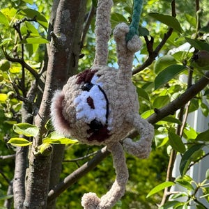 May include: A crocheted sloth toy made of light tan yarn with dark brown eye patches and a white face mask. The plush figure has long limbs and is suspended by a green yarn cord, featuring black button eyes and a small dark brown nose.