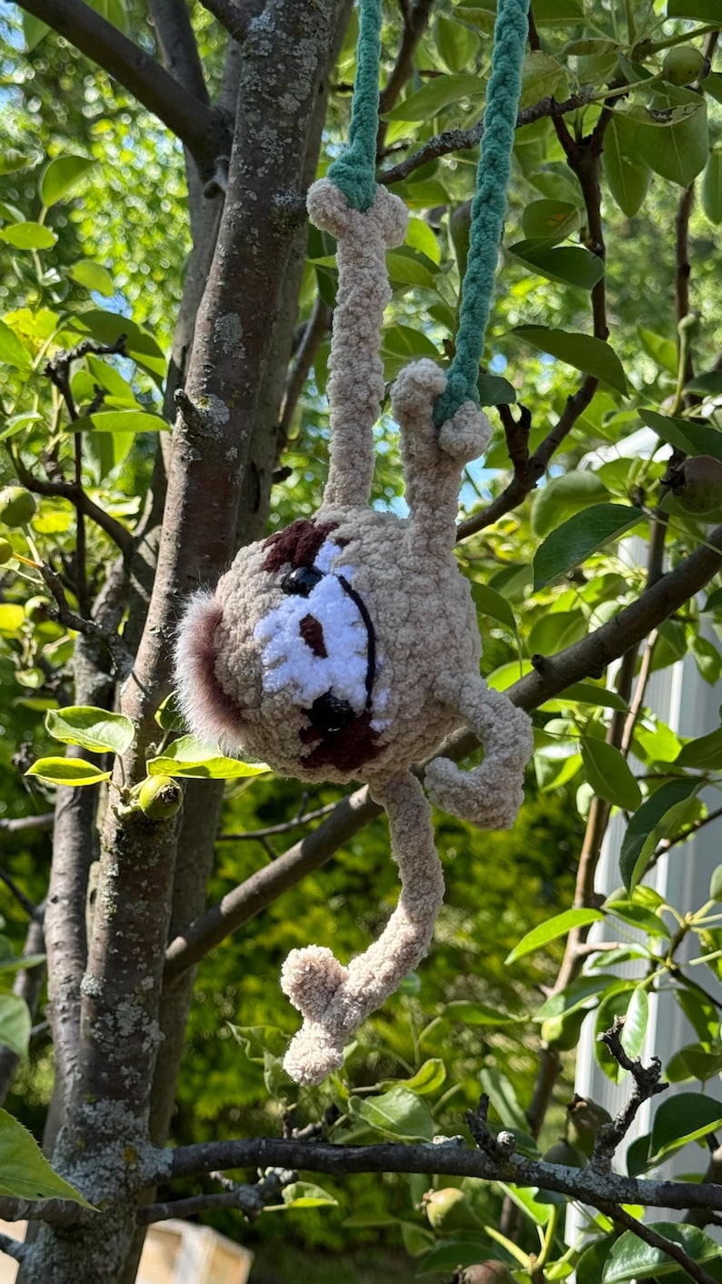 May include: A crocheted sloth toy made of light tan yarn with dark brown eye patches and a white face mask. The plush figure has long limbs and is suspended by a green yarn cord, featuring black button eyes and a small dark brown nose.