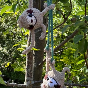 May include: Two crocheted sloth figures in light beige yarn with dark brown eye patches and white facial details. Each sloth hangs from a long, braided green yarn cord, featuring long limbs and a soft, fuzzy texture, suspended from tree branches.