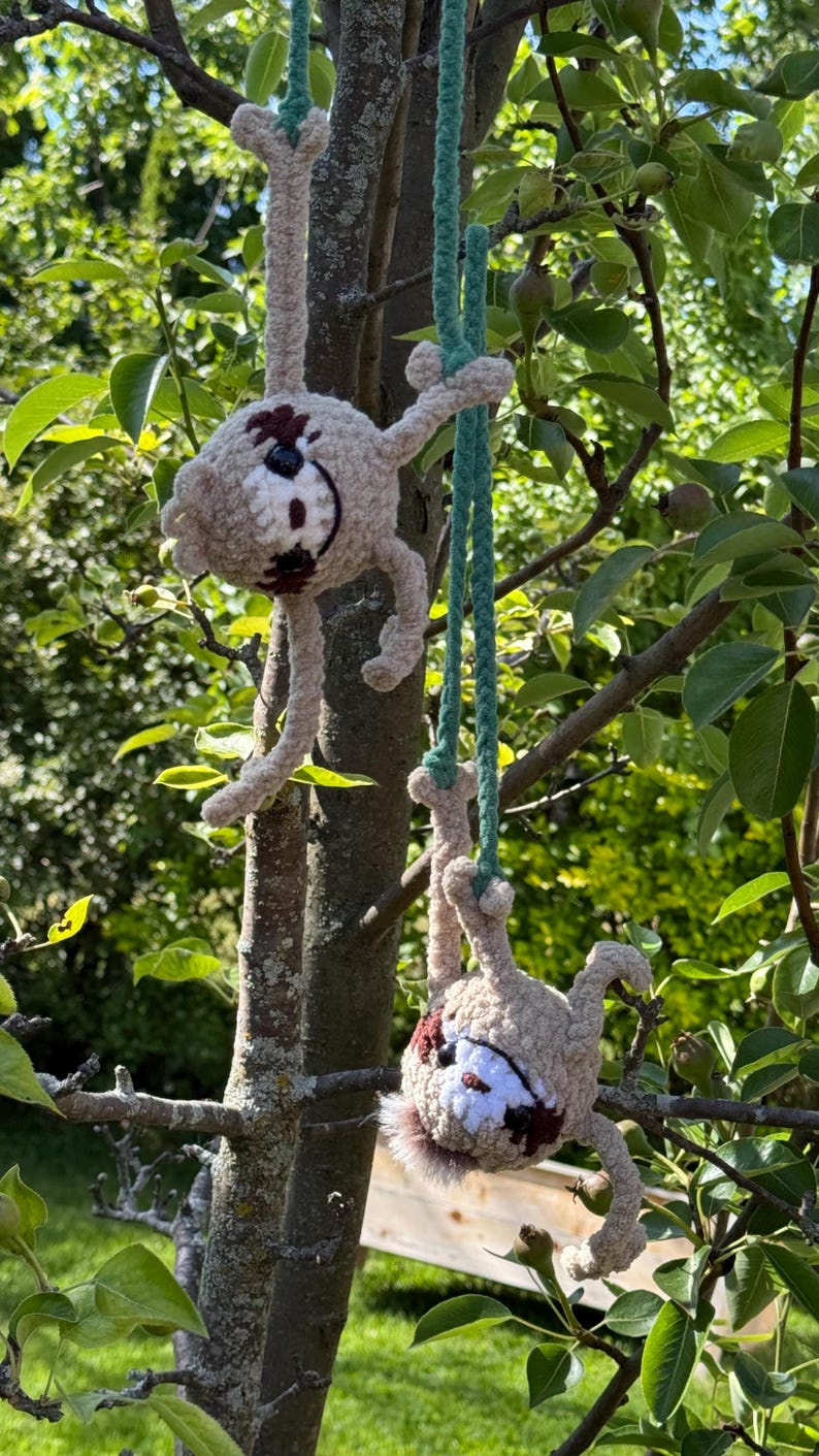 May include: Two crocheted sloth figures in light beige yarn with dark brown eye patches and white facial details. Each sloth hangs from a long, braided green yarn cord, featuring long limbs and a soft, fuzzy texture, suspended from tree branches.