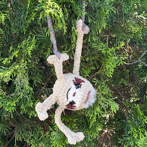 May include: A beige crocheted sloth plush toy with dark brown eye patches and a small tuft of fur on its head. The sloth has long limbs and is suspended by a light purple yarn loop, featuring black plastic safety eyes and a stitched mouth.