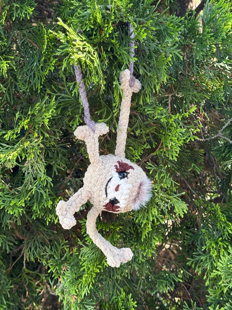 May include: A beige crocheted sloth plush toy with dark brown eye patches and a small tuft of fur on its head. The sloth has long limbs and is suspended by a light purple yarn loop, featuring black plastic safety eyes and a stitched mouth.