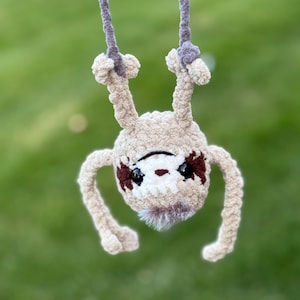 May include: A crocheted sloth ornament made from beige yarn with dark brown eye patches and black plastic eyes. The sloth is hanging upside down by two grey yarn loops attached to its feet. It features a small tuft of white faux fur at the chin.
