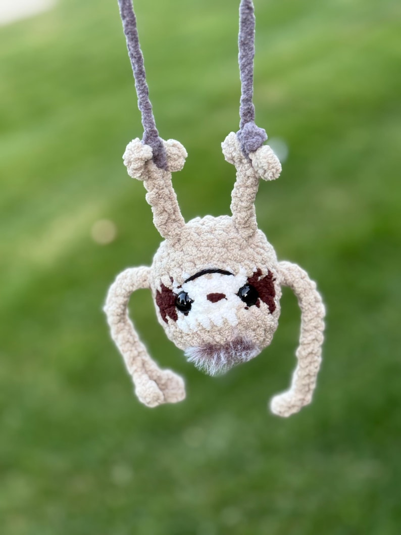 May include: A crocheted sloth ornament made from beige yarn with dark brown eye patches and black plastic eyes. The sloth is hanging upside down by two grey yarn loops attached to its feet. It features a small tuft of white faux fur at the chin.