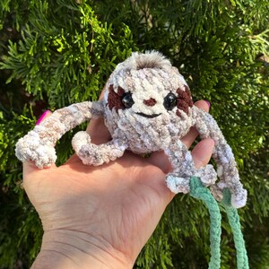 May include: A small, round, crocheted sloth plush toy made of variegated beige, brown, and white yarn. It features dark brown eye patches, black safety eyes, a small embroidered nose, and a tuft of faux fur on top, with long arms and light green braided cords.