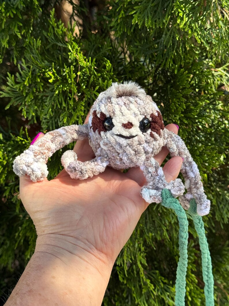 May include: A small, round, crocheted sloth plush toy made of variegated beige, brown, and white yarn. It features dark brown eye patches, black safety eyes, a small embroidered nose, and a tuft of faux fur on top, with long arms and light green braided cords.